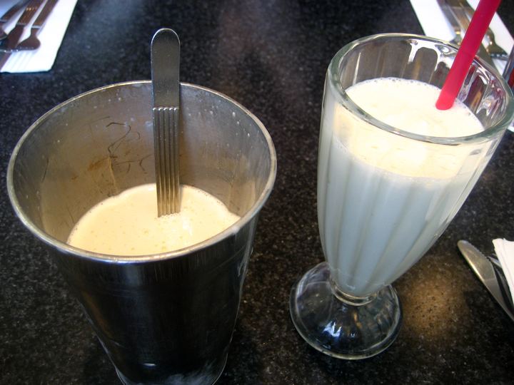 A tall milkshake glass with a metal mixing cup beside it